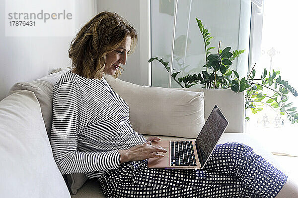 Frau sitzt zu Hause auf dem Sofa und benutzt Laptop im Wohnzimmer