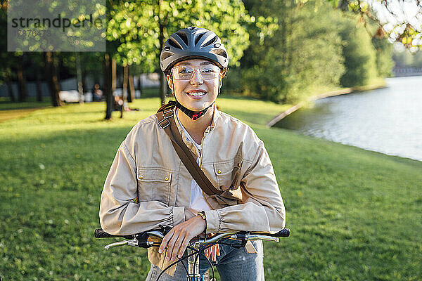 Lächelnde Frau mit Helm sitzt auf dem Fahrrad im Park