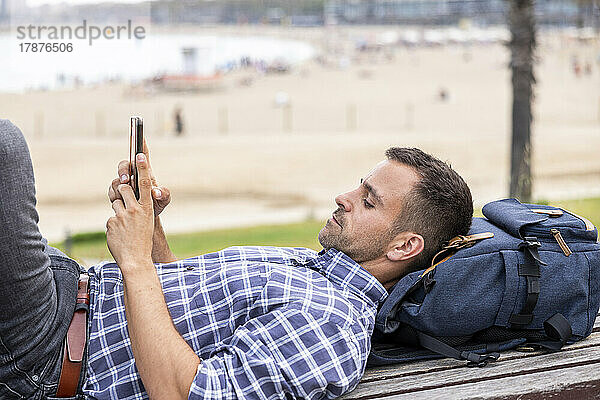 Reifer Mann liegt auf Bank und benutzt Mobiltelefon am Strand