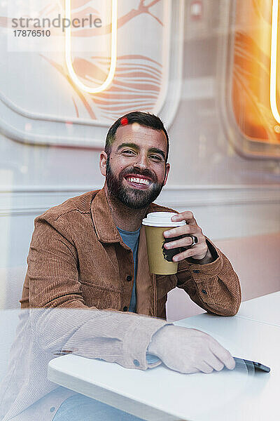 Fröhlicher junger Mann mit Einwegkaffeetasse sitzt am Tisch im Café
