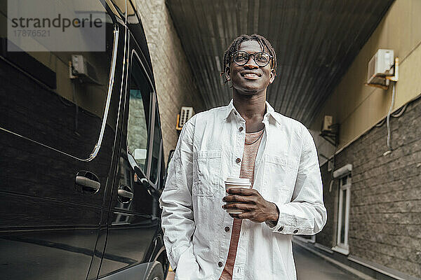 Lächelnder junger Mann mit Einwegkaffeetasse steht neben dem Lieferwagen