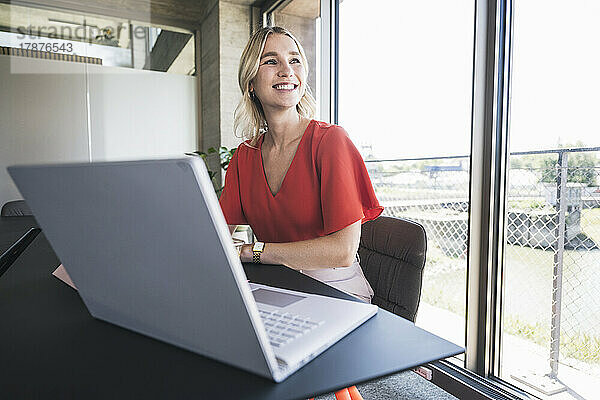 Glückliche blonde Geschäftsfrau sitzt mit Laptop am Schreibtisch im Büro