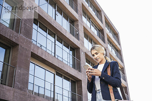 Lächelnde junge Frau benutzt Smartphone vor Gebäude