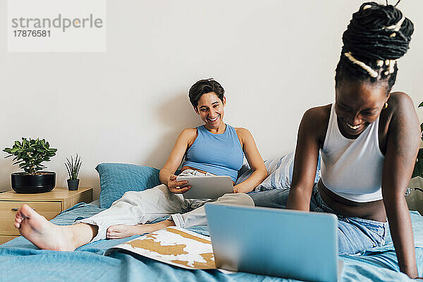 Happy woman using laptop with girlfriend in background on bed at home