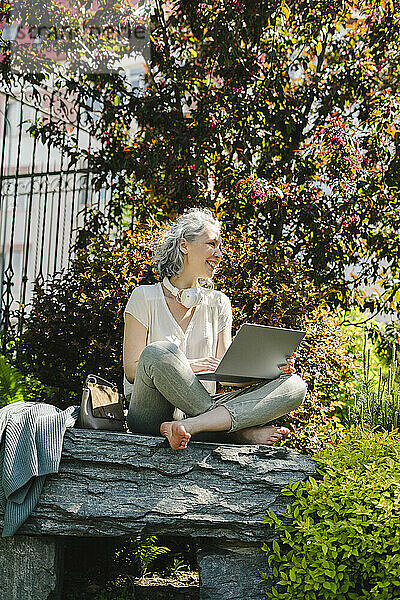 Lächelnder Freiberufler sitzt mit Laptop auf einem Felsen im Park