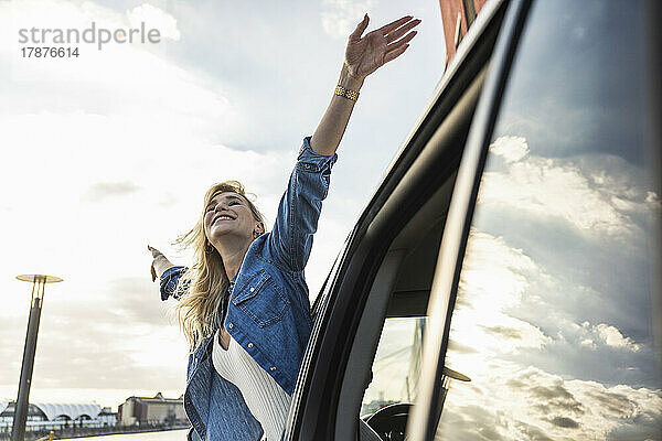 Carefree young woman with arms outstretched leaning out of car window
