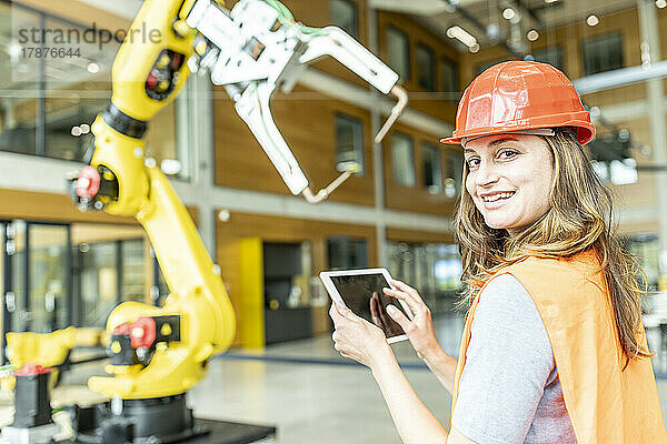 Lächelnde junge Arbeiterin  die den Roboterarm mit einem digitalen Tablet steuert