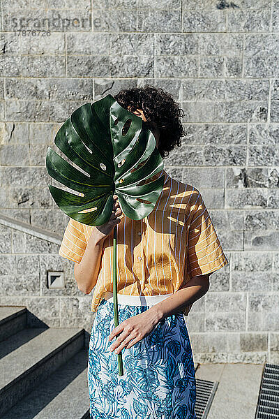 Frau bedeckt Gesicht an sonnigem Tag mit Monstera-Blatt