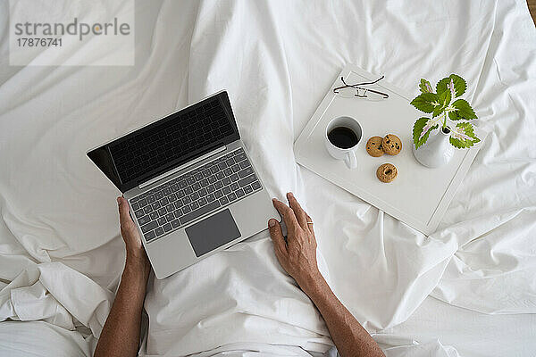Hände eines Mannes mit Laptop und Frühstück auf dem Bett