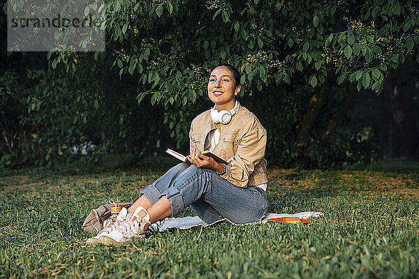 Lächelnde Frau sitzt mit Buch und kabellosen Kopfhörern im Park