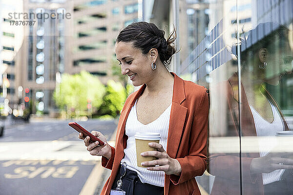 Lächelnde Geschäftsfrau hält Einwegbecher und Smartphone an Glaswand gelehnt