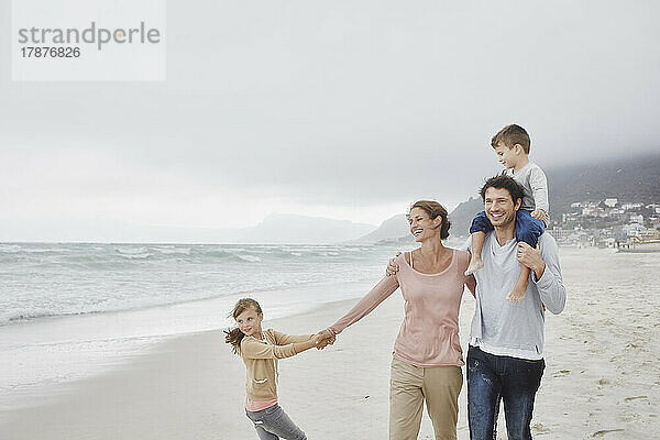 Eltern gehen mit zwei Kindern am Strand spazieren und tragen ihren Sohn auf den Schultern