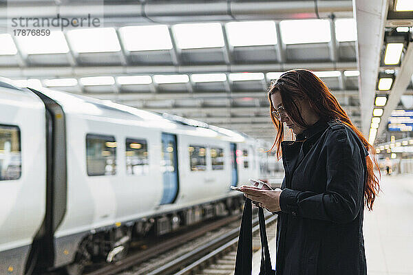 Rothaarige Frau benutzt Smartphone am Bahnsteig