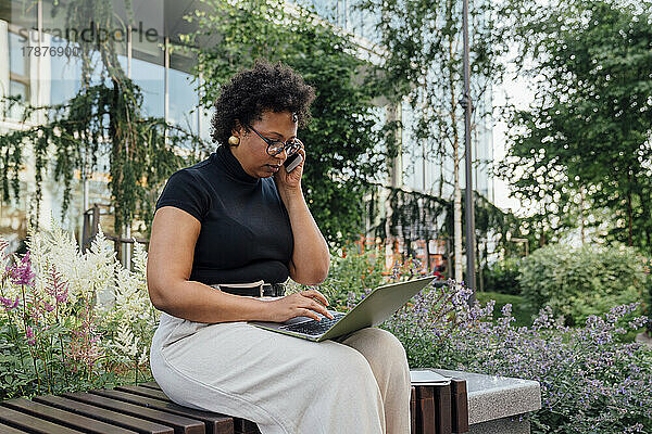 Businesswoman using laptop and talking on mobile phone sitting on bench at office park