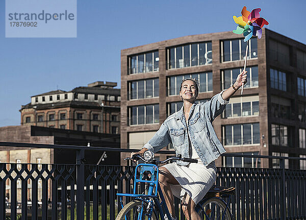 Glückliche Frau mit Windradspielzeug  die an einem sonnigen Tag Fahrrad fährt