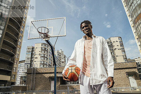 Lächelnder Mann  junger Mann  der mit Basketball auf dem Sportplatz steht