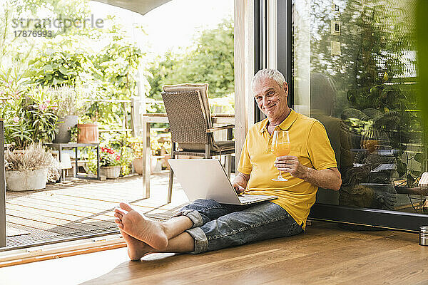 Lächelnder Mann hält zu Hause ein Glas Saft mit Laptop in der Hand