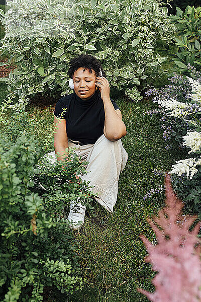 Young woman with eyes closed listening music through wireless headphones sitting amidst plants at park