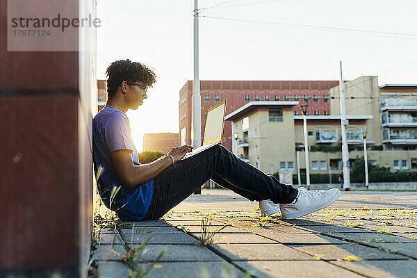 Junger Mann lernt am Laptop und sitzt bei Sonnenuntergang auf dem Fußweg in der Stadt