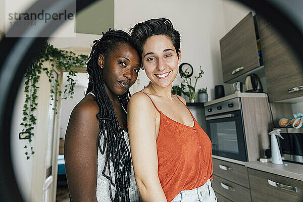 Smiling lesbian couple standing at home seen through ring light