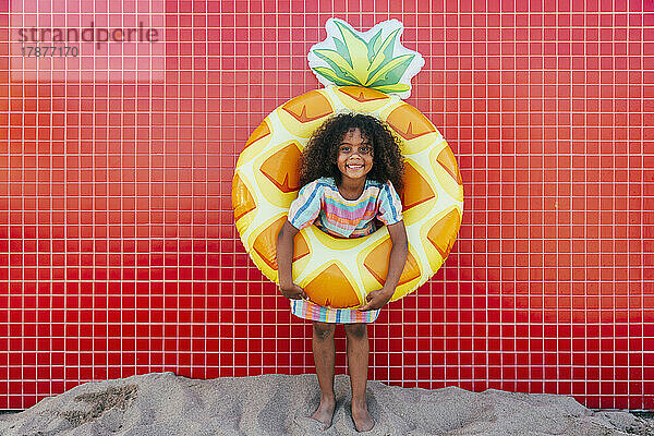 Lächelndes Mädchen mit Schwimmkörper  das vor einer Mauer am Strand steht