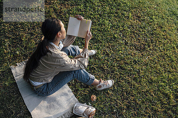 Frau liest Buch mit Essen und sitzt auf Gras im Park