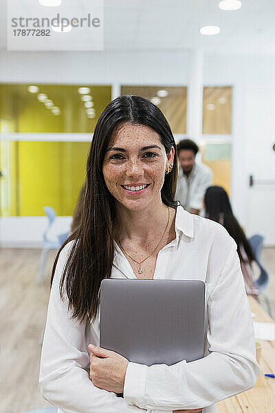 Porträt einer lächelnden Geschäftsfrau mit Laptop im Büro und Kollegen im Hintergrund