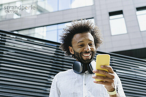 Glücklicher Mann mit Kopfhörern und Smartphone vor dem Gebäude