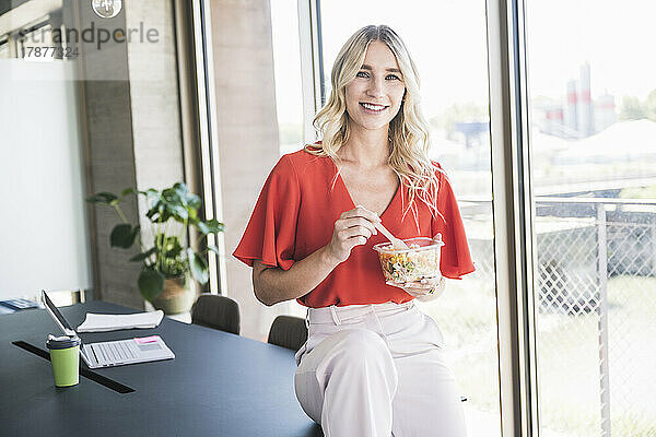 Lächelnde Geschäftsfrau hält eine Lunchbox in der Hand und sitzt auf dem Schreibtisch im Büro