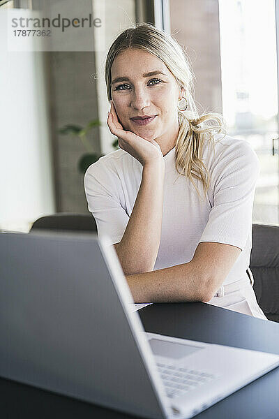 Blonde Geschäftsfrau stützt sich auf den Ellenbogen und sitzt am Schreibtisch im Büro