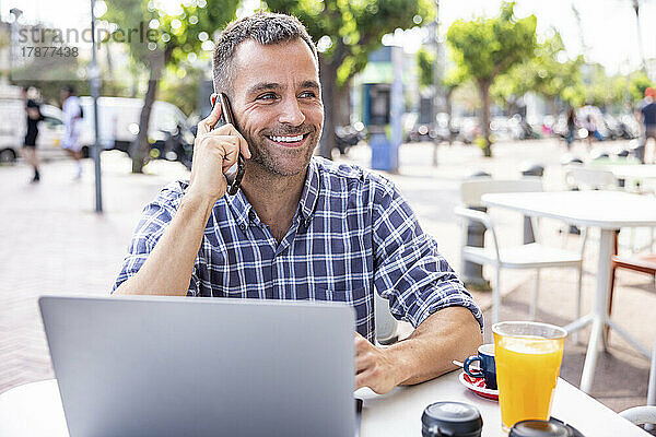Lächelnder reifer Freiberufler  der über sein Smartphone spricht und mit einem Laptop im Straßencafé sitzt