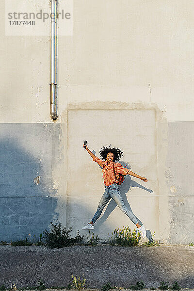 Glückliche Frau mit Smartphone springt vor Wand