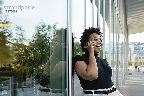 Glückliche Geschäftsfrau spricht mit Smartphone und lehnt an Glaswand