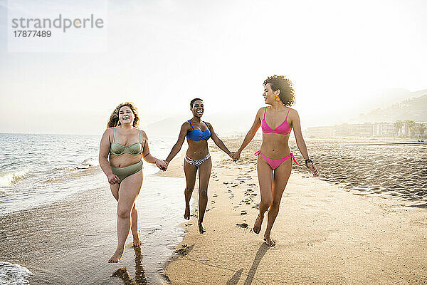Fröhliche Freunde halten sich an den Händen und laufen an einem sonnigen Tag am Strand