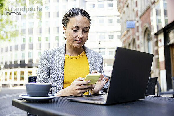 Geschäftsfrau sitzt mit Laptop im Straßencafé und benutzt Smartphone