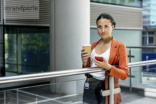 Geschäftsfrau hält Einwegbecher und Smartphone am Geländer