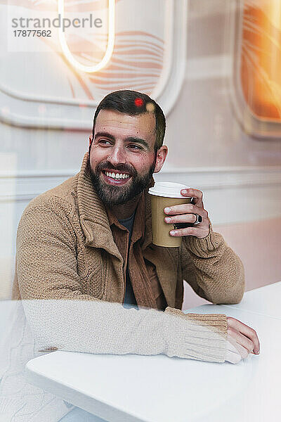 Happy young man with disposable coffee cup sitting at table in cafe