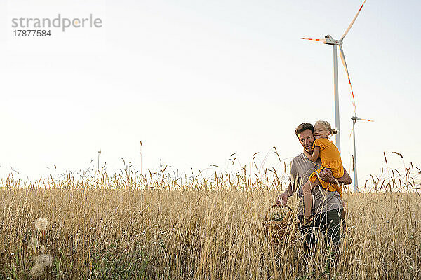 Vater trägt Tochter auf Weizenfeld mit Windkraftanlagen