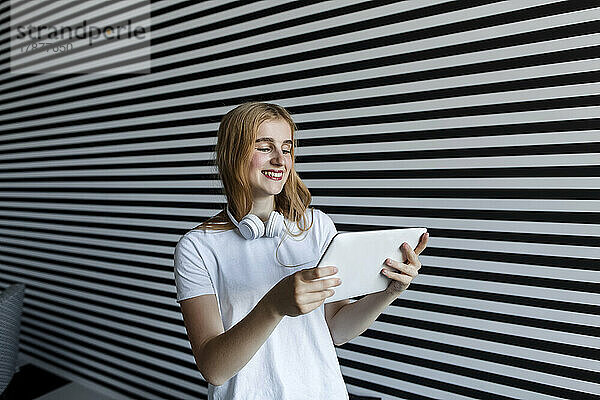 Lächelndes schönes Mädchen mit Kopfhörern  das Tablet-PC an der gestreiften Wand beobachtet
