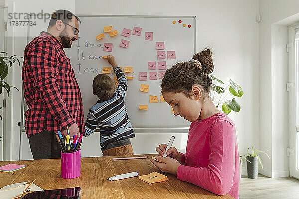 Mädchen schreibt mit Filzstift  während Bruder zu Hause Klebezettel auf Whiteboard klebt