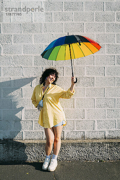 Lächelnde Frau mit Regenbogenschirm posiert vor der Wand