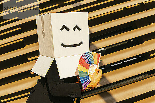 Geschäftsmann trägt Schachtel mit Smiley und hält an einem sonnigen Tag einen regenbogenfarbenen Handfächer