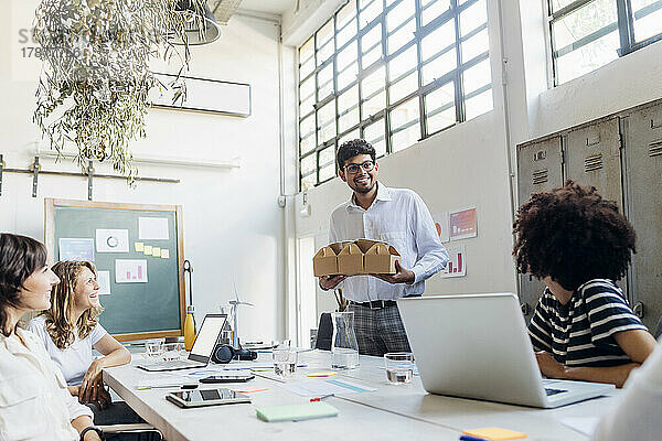 Lächelnder Geschäftsmann mit Lunchbox und Blick auf Kollegen im Büro