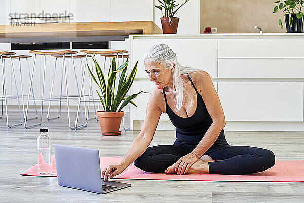 Fitte Frau sitzt zu Hause mit Laptop auf einer Trainingsmatte
