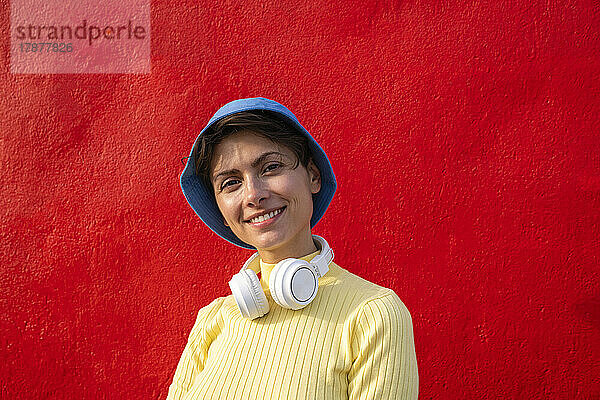 Lächelnde Frau mit Kopfhörern und Fischerhut vor roter Wand