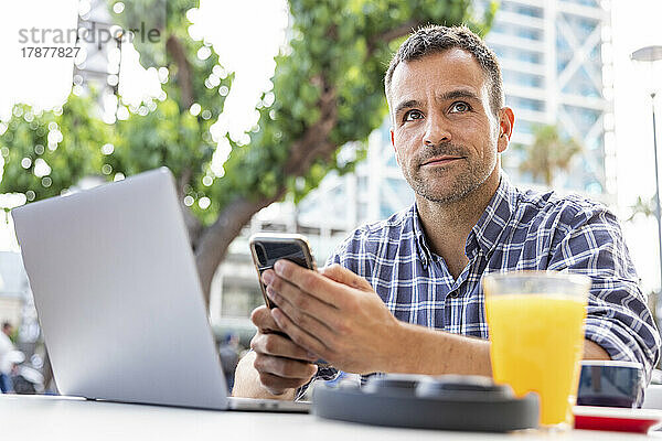 Reifer Mann sitzt mit Laptop und Smartphone im Straßencafé