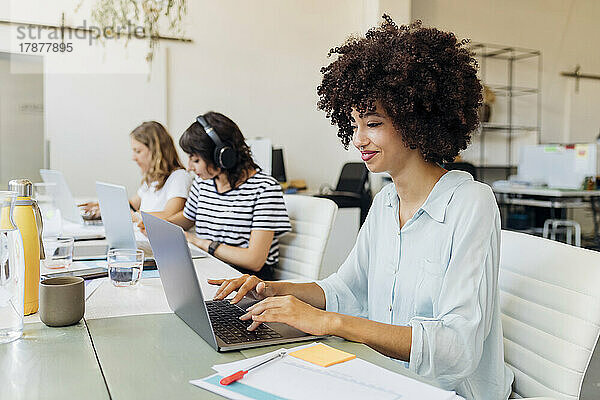 Lächelnde Geschäftsfrau mit lockigem Haar  die im Büro am Laptop arbeitet