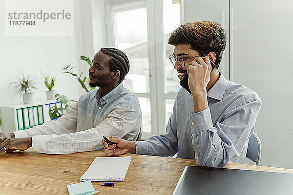 Glücklicher Geschäftsmann  der am Handy sitzt und neben einem Kollegen im Büro sitzt