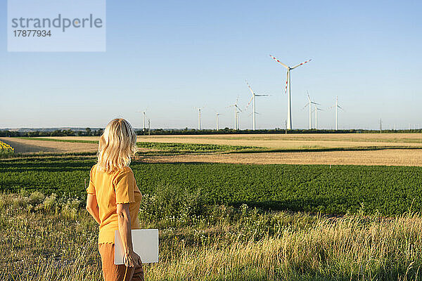 Frau hält Laptop und blickt auf Windkraftanlage im Feld