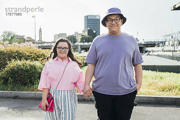 Lächelnder Bruder hält die Hand seiner Schwester mit Down-Syndrom  die auf dem Fußweg steht